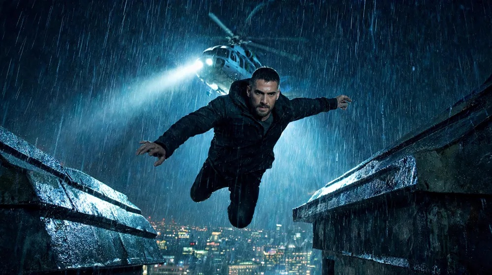 A man leaping between rooftops in the rain as a helicopter searchlight cuts the night